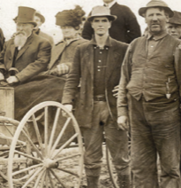 A sepia-toned photograph of two men standing in front of a carriage. The photo is focused on the man in the middle, wearing a button-down, jacket, and hat. This is assumed to be James Wilson.